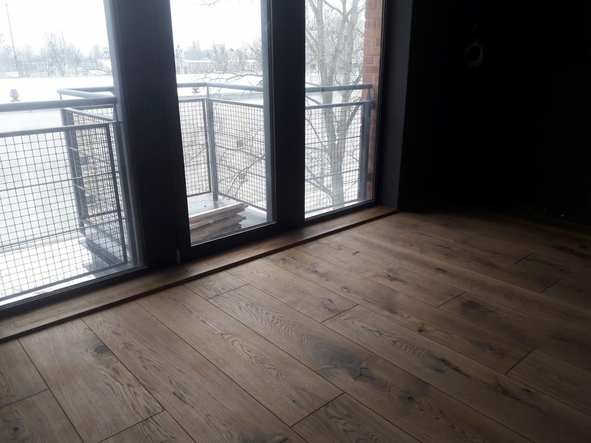 Ułożony parkiet z desek w odcieniach brązu i beżu, widok na balkon z metalową balustradą i zimowy krajobraz za oknem, czarna ściana w tle.