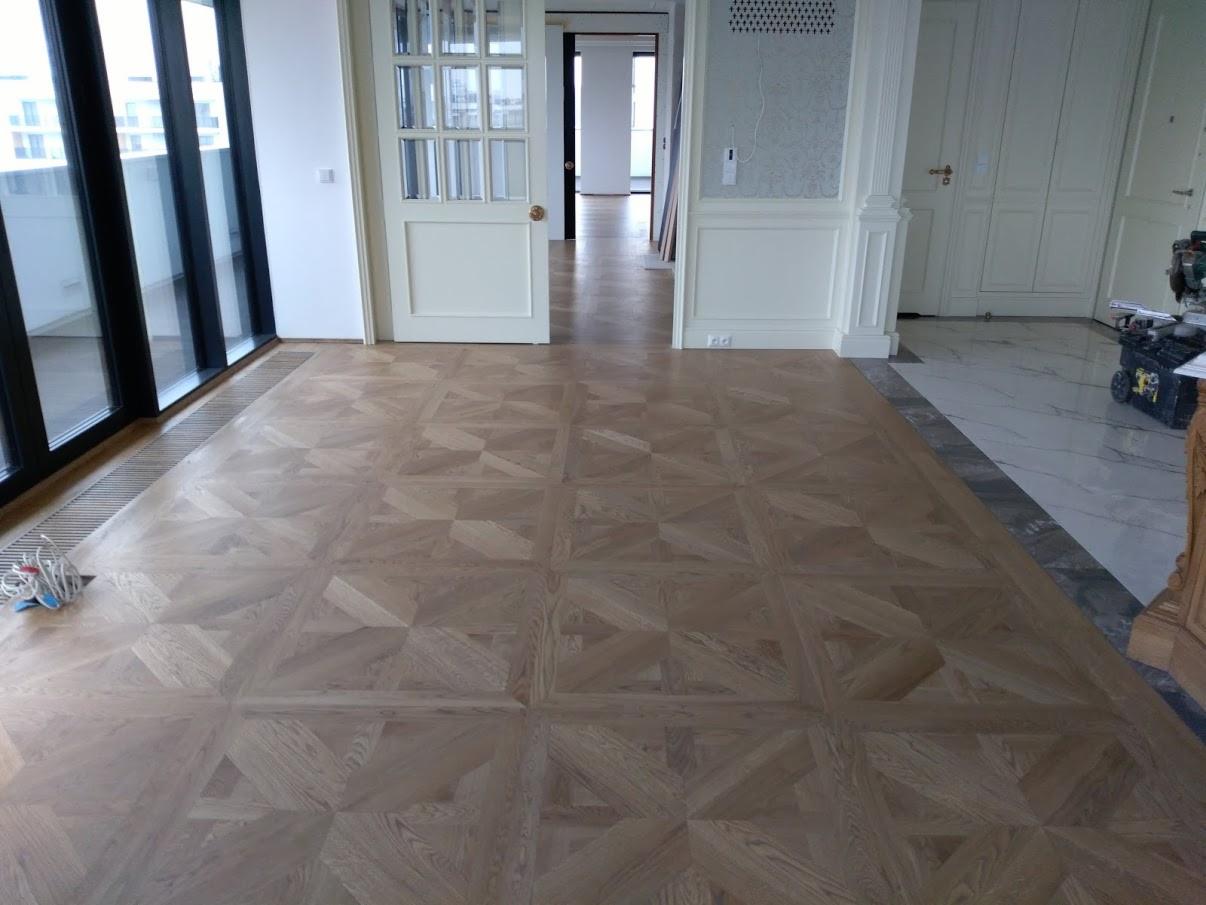 Wzorzysta drewniana podłoga z parkietu ułożona w kwadraty, widok na przestronne wnętrze z dużymi oknami i białymi drzwiami.