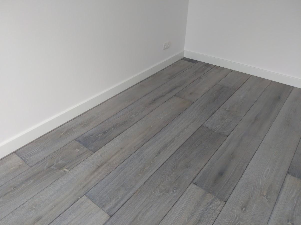 Szare panele podłogowe ułożone w rogu pokoju z białymi ścianami i listwami przypodłogowymi.