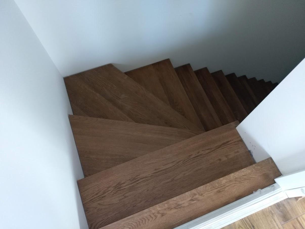 Widok z góry na drewniane schody z widoczną strukturą drewna, umieszczone w jasnym, minimalistycznym wnętrzu z białymi ścianami.