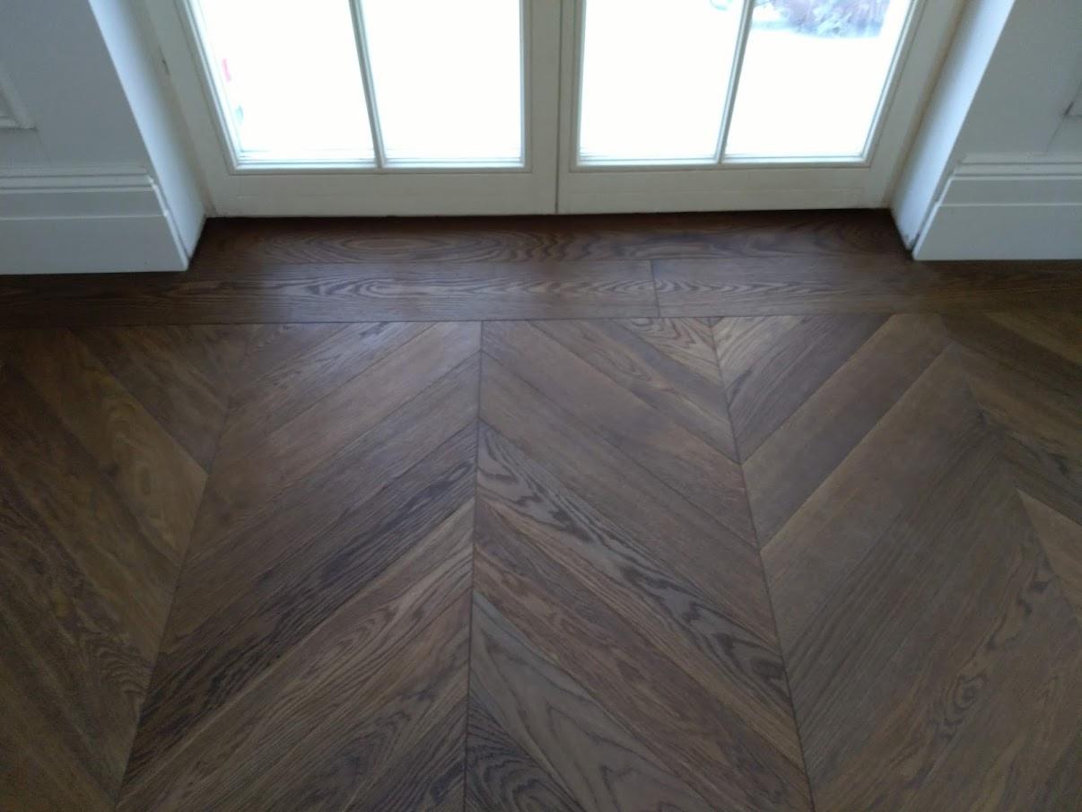 Ułożony parkiet w jodełkę francuską, ciemne drewno, widoczne okno z białymi ramami i listwami przypodłogowymi.