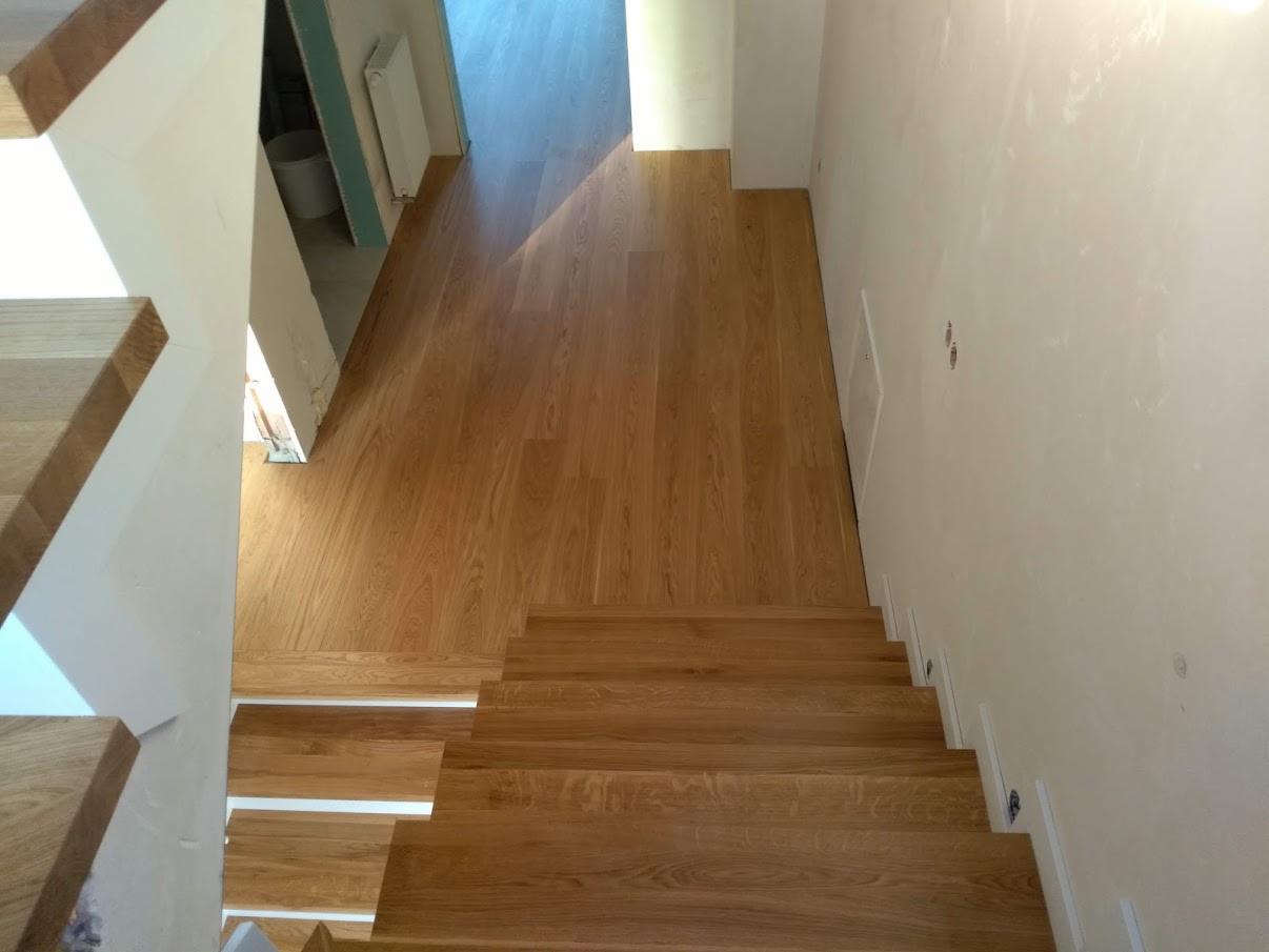 Widok z góry na drewniane schody łączące się z parkietem w jasnym odcieniu, widoczne białe listwy przypodłogowe i fragment korytarza w tle.