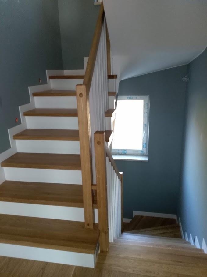 Drewniane schody z białymi podstopnicami i drewnianą balustradą, oświetlone przez okno, prowadzące na dół.