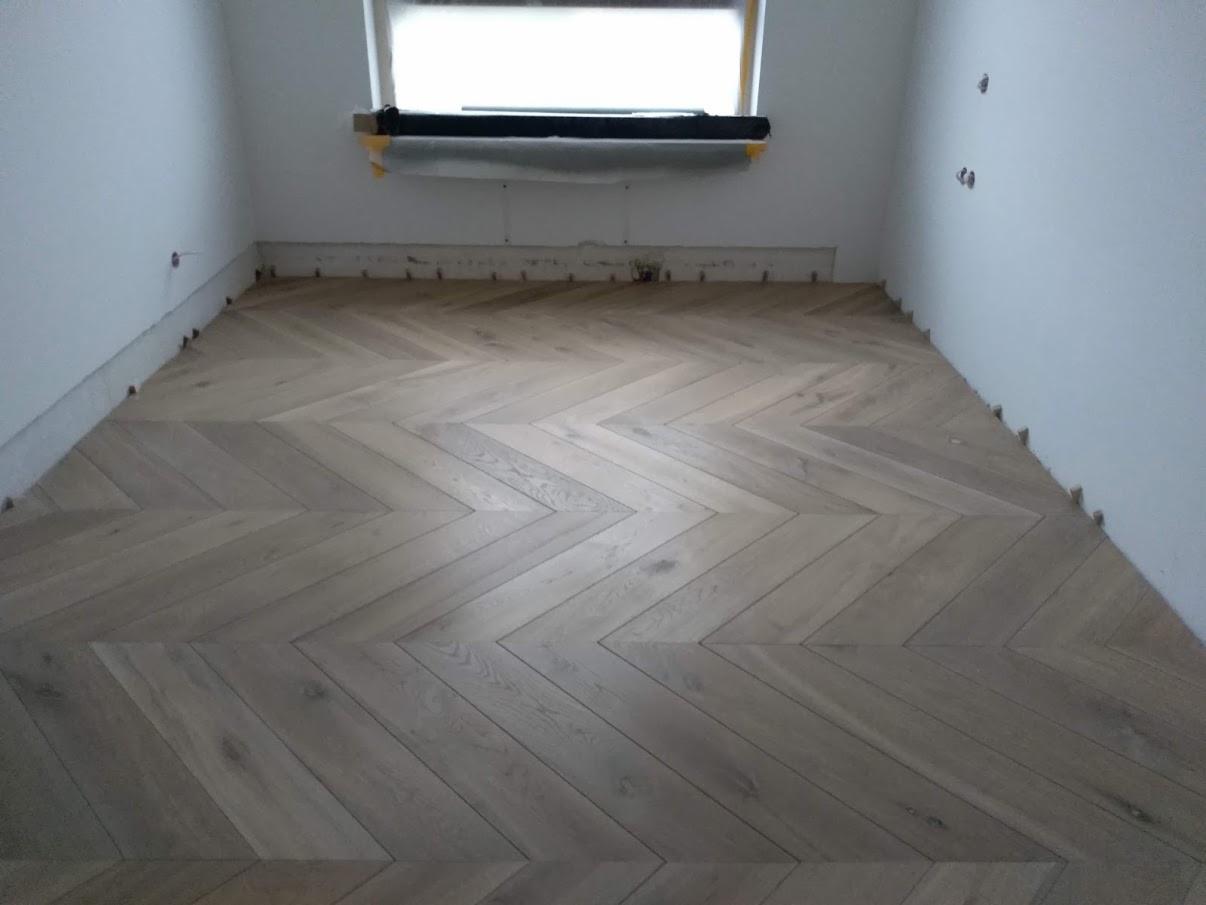 Świeżo ułożona podłoga z parkietu w jodełkę, widoczne kliny dystansowe przy ścianach, pokój w trakcie wykańczania.