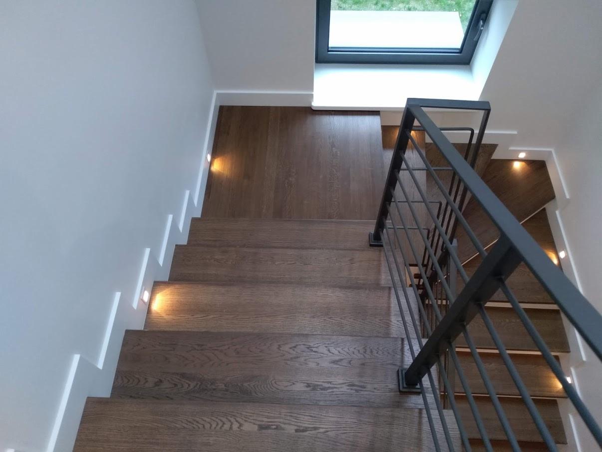 Nowoczesne schody drewniane z metalową balustradą, oświetleniem punktowym w ścianie i pod stopniami, widok z góry na pierwsze stopnie.