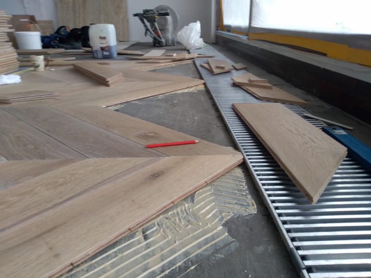 Fragment podłogi z jasnych desek układanych w jodełkę, widoczny czerwony ołówek, klej i narzędzia w tle, proces montażu.