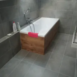 Wykończona łazienka na poddaszu ze skośnym oknem dachowym, szare płytki na podłodze i ścianach, obudowana drewnopodobną płytą wanna z czerwoną ściereczką, widoczne butelki z kosmetykami.