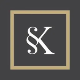 Logo kancelarii prawnej: stylizowany paragraf i litera K w kolorze białym na ciemnoszarym tle, obramowane złotą ramką.