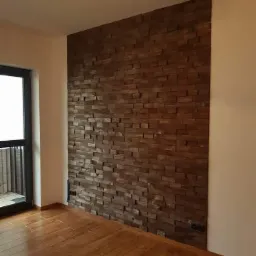 Ściana akcentowa z drewnianych paneli 3D w nowoczesnym wnętrzu, z widoczną podłogą z paneli i oknem balkonowym po lewej stronie.