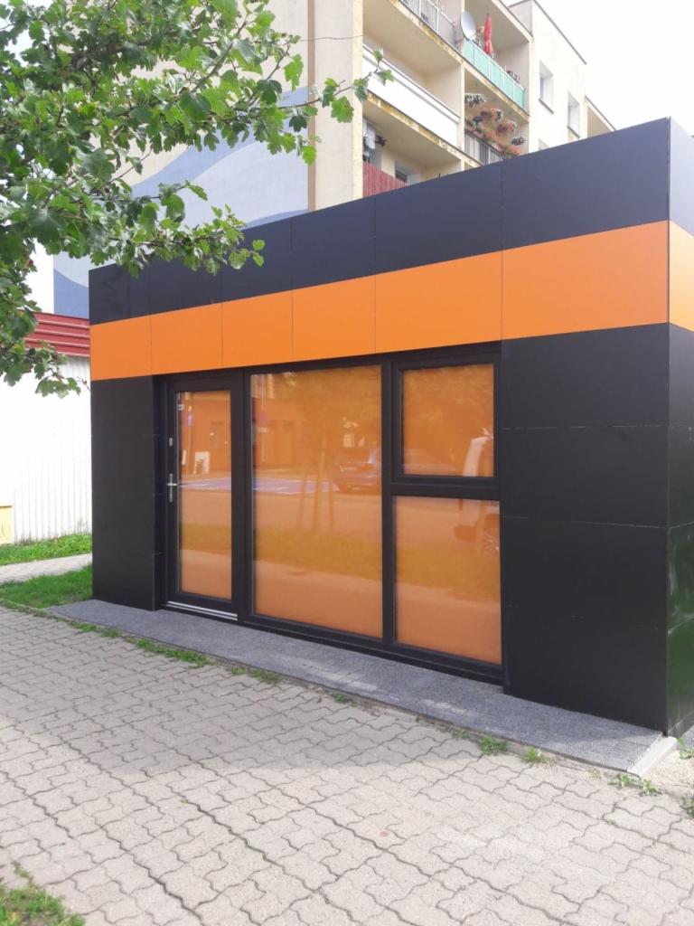 Wykończony pawilon handlowy w Sławnie z czarnymi ścianami i pomarańczowym pasem, widoczne przeszklone drzwi i okna.