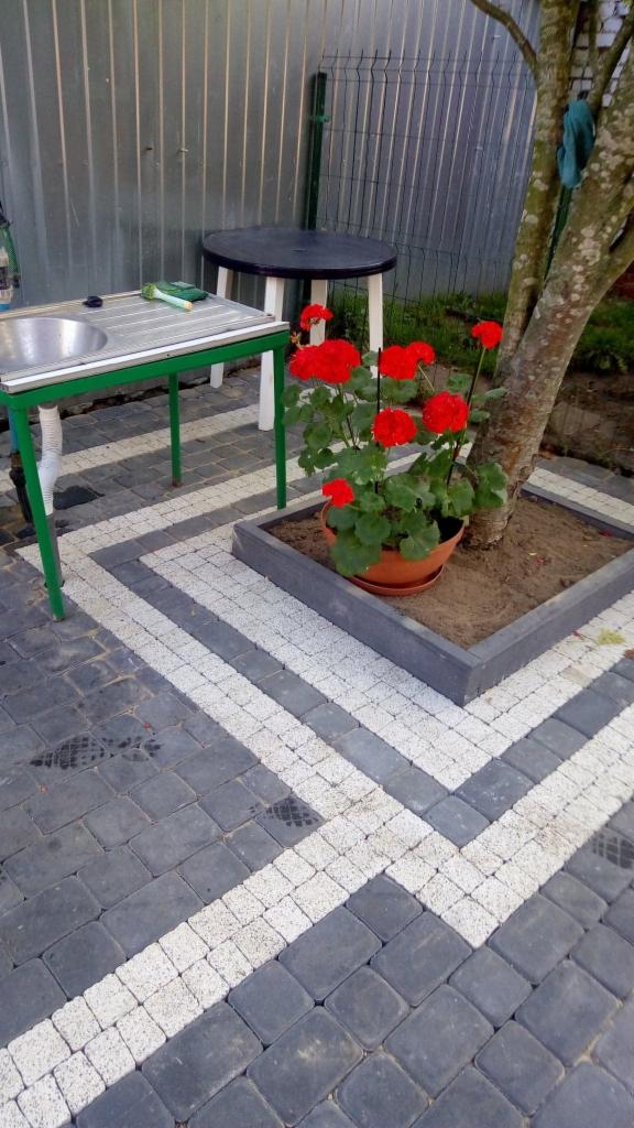 Ułożona kostka brukowa w szarych i białych pasach, z donicą czerwonych kwiatów przy pniu drzewa, obok stolik i umywalka na zewnątrz.