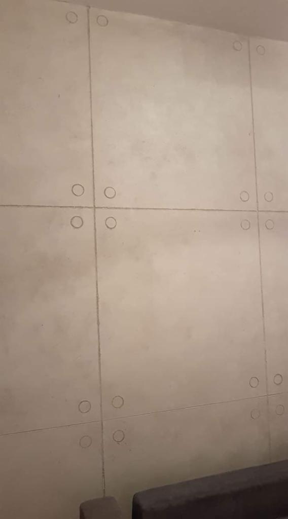 Ściana z szarych płyt imitujących beton, z widocznymi okrągłymi wgłębieniami w rogach każdej płyty, tworzącymi regularny wzór.