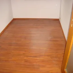 Pokój z nowo ułożoną podłogą z paneli o ciepłym, brązowym odcieniu drewna, widoczne listwy przypodłogowe i fragment drzwi z szybą.