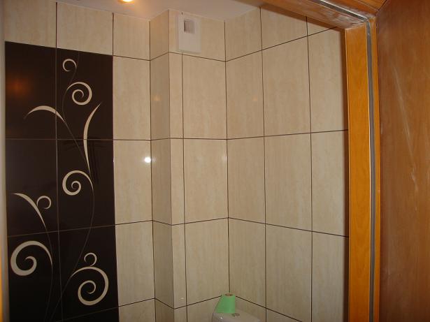 Wykończona łazienka z beżowymi płytkami i dekoracyjną czarną ścianą z motywem roślinnym, widoczny fragment toalety z zielonym papierem toaletowym.