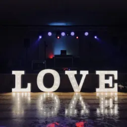 Podświetlany napis LOVE na scenie z parkietem odbijającym światła, w tle sprzęt DJ-ski i oświetlenie sceniczne.