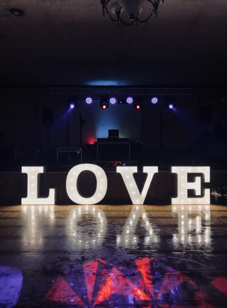 Podświetlany napis LOVE na scenie z parkietem odbijającym światła, w tle sprzęt DJ-ski i oświetlenie sceniczne.
