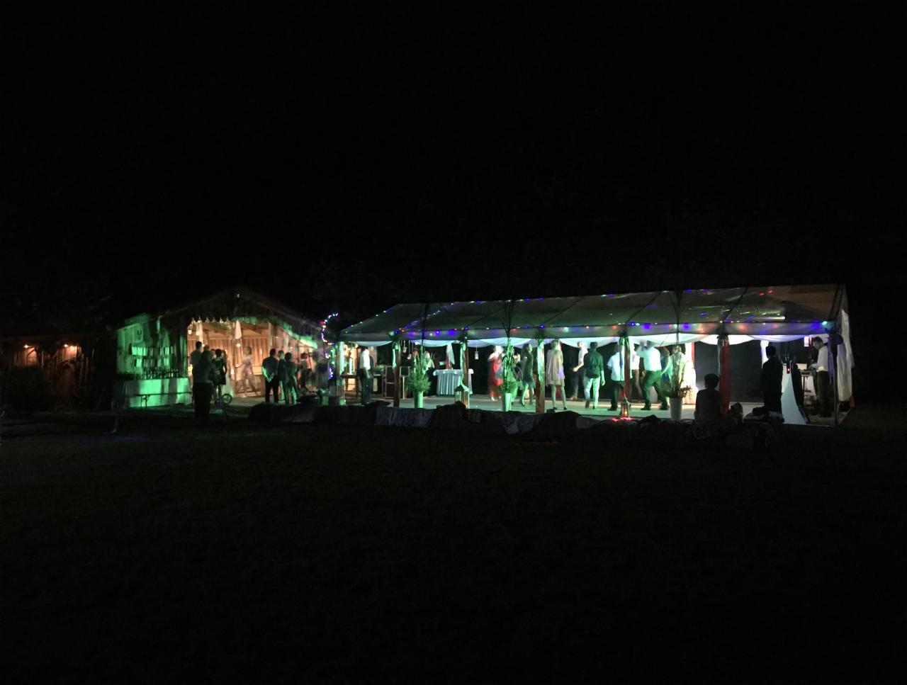 Zabawa taneczna pod oświetlonym namiotem w nocy, z budynkiem w tle oświetlonym na zielono, uczestnicy w ruchu.