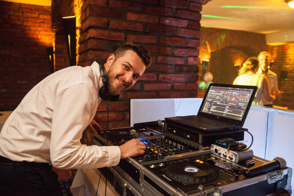 Uśmiechnięty DJ w białej koszuli i słuchawkach, obsługujący mikser i laptop na tle ceglanej ściany i tańczącej pary w tle.