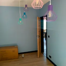 Wnętrze pokoju z błękitnymi ścianami i drewnianą podłogą, oświetlone wiszącymi lampami w kolorach r&oacute;żu, fioletu i turkusu, z otwartymi drzwiami i kartonem w rogu.