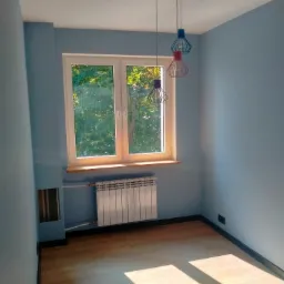 Pomalowane na niebiesko wnętrze pokoju z drewnianą podłogą, białym grzejnikiem pod oknem z widokiem na zieleń i wiszącą lampą z trzema kolorowymi kloszami w kształcie klatek.