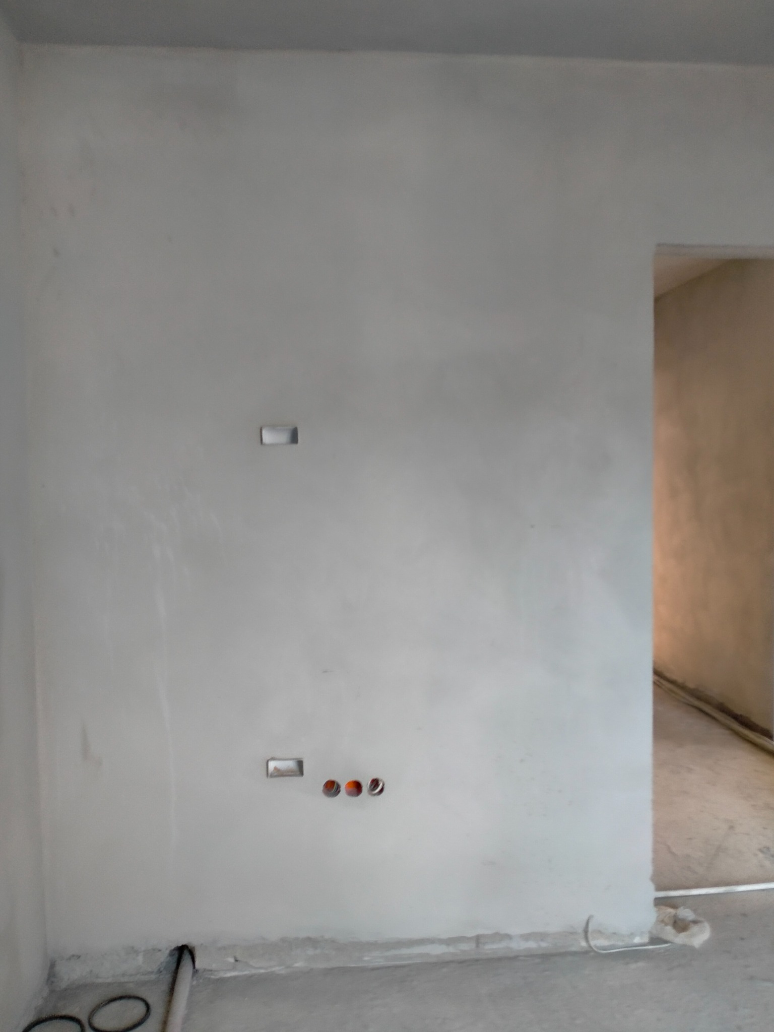 Świeżo położony tynk maszynowy na ścianie z widocznymi puszkami elektrycznymi i przewodami w podłodze, przygotowanie do dalszych prac wykończeniowych.