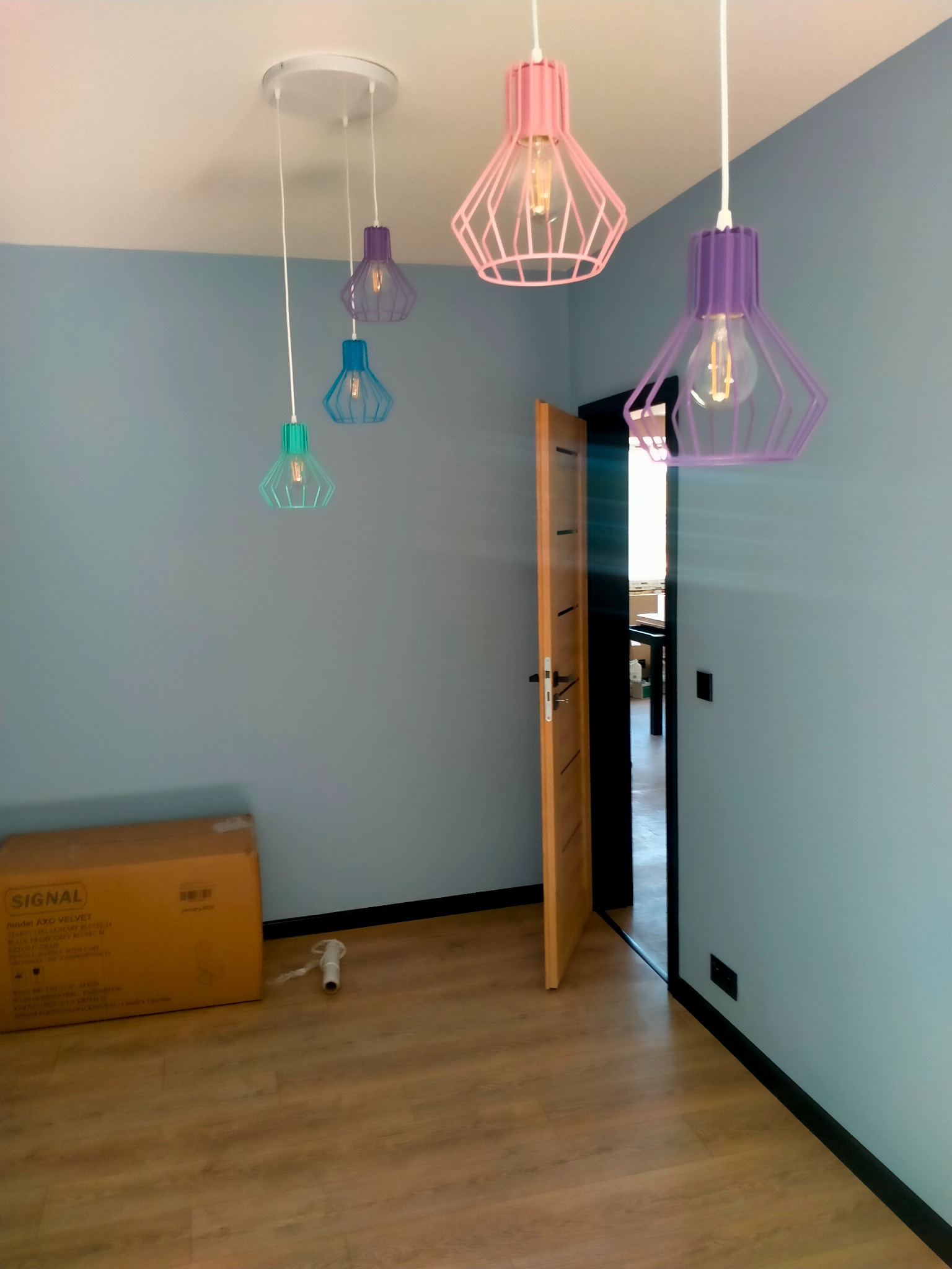 Wnętrze pokoju z błękitnymi ścianami i drewnianą podłogą, oświetlone wiszącymi lampami w kolorach różu, fioletu i turkusu, z otwartymi drzwiami i kartonem w rogu.