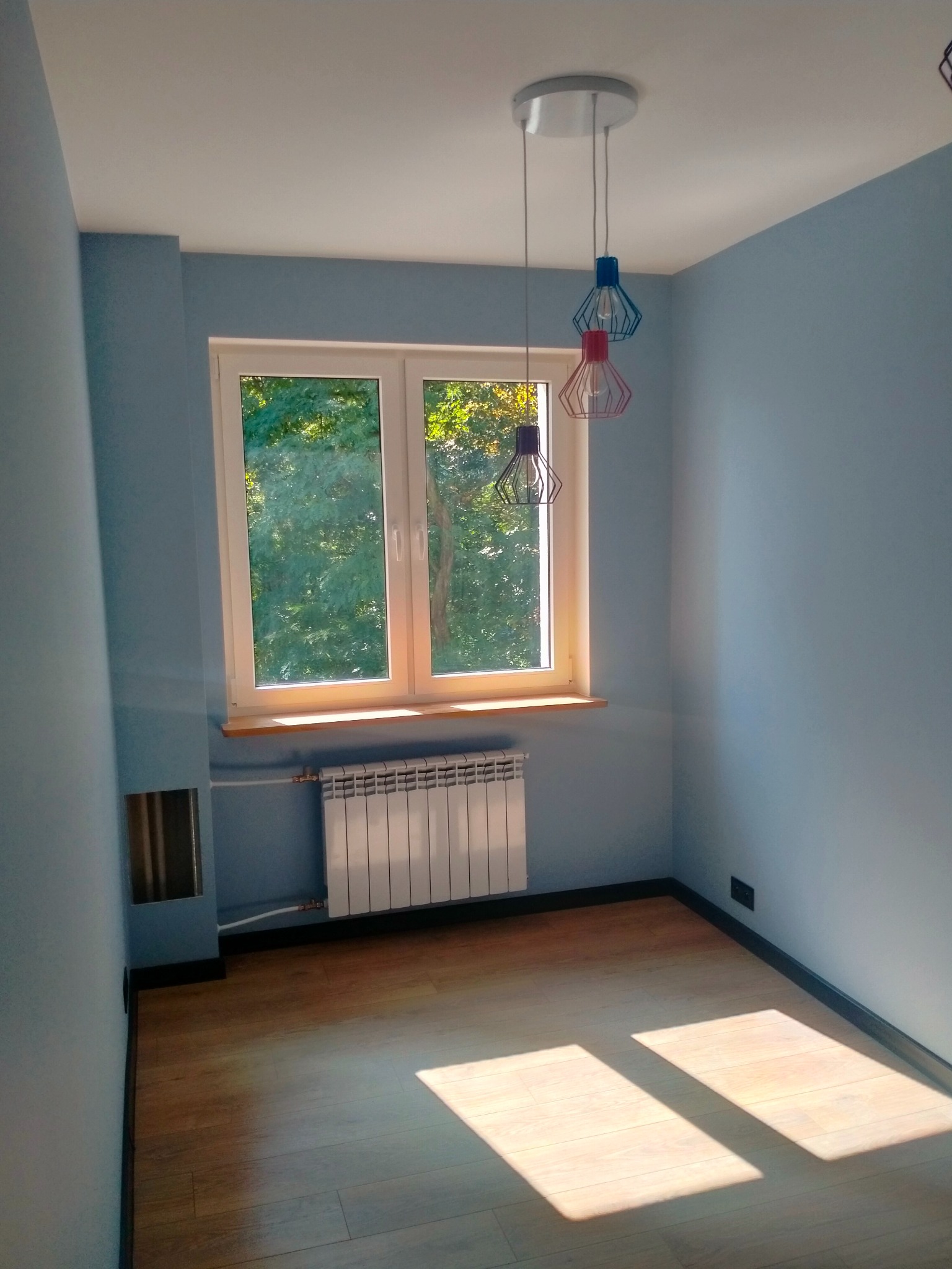 Pomalowane na niebiesko wnętrze pokoju z drewnianą podłogą, białym grzejnikiem pod oknem z widokiem na zieleń i wiszącą lampą z trzema kolorowymi kloszami w kształcie klatek.