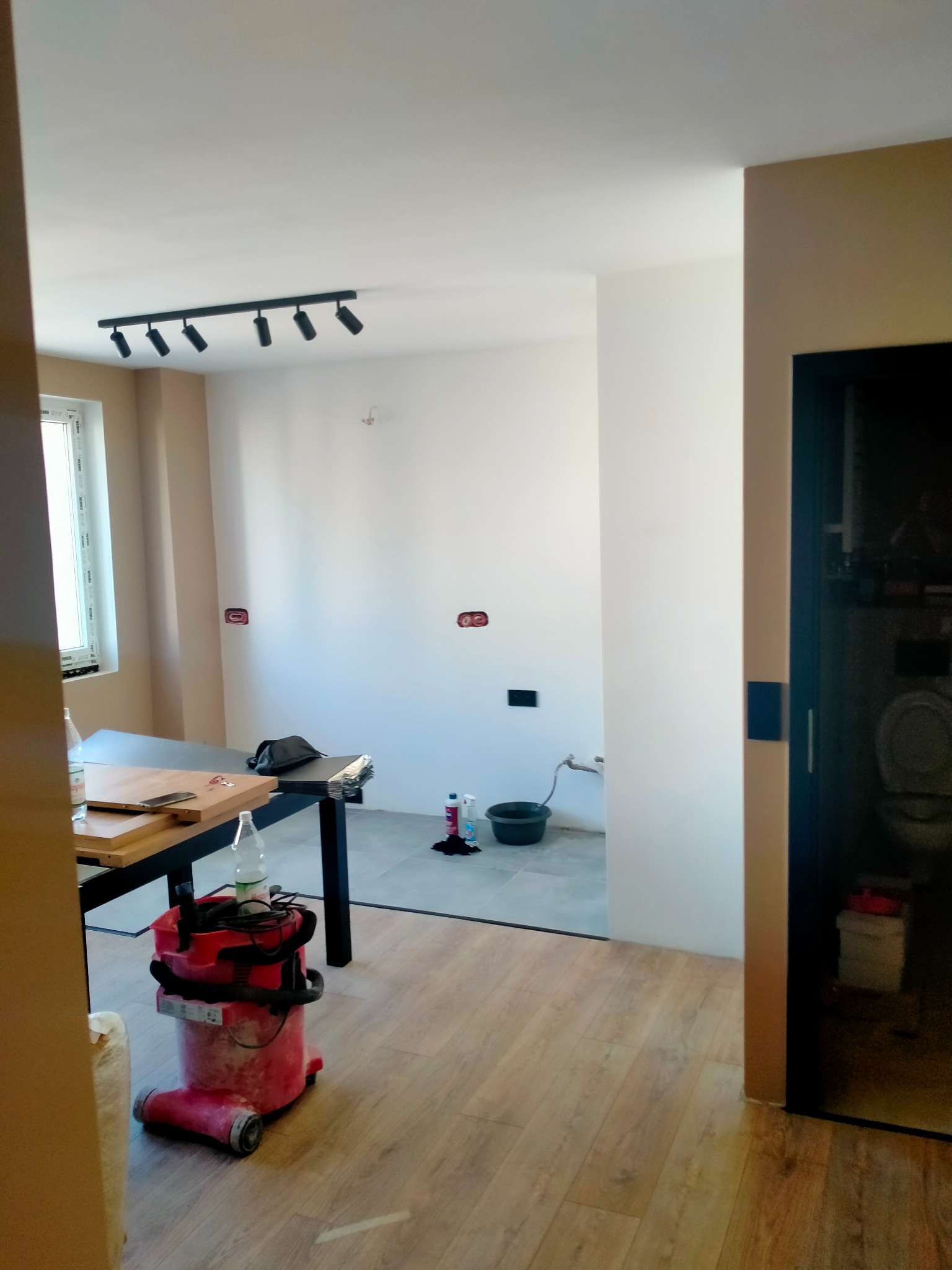 Wnętrze w trakcie remontu: widoczne surowe ściany z puszkami elektrycznymi, podłoga częściowo wyłożona płytkami, częściowo panelami, czerwony odkurzacz przemysłowy, stół roboczy z narzędziami...