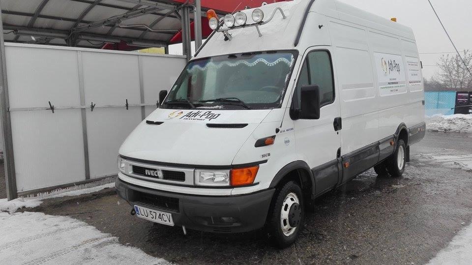 Biały bus marki Iveco z logo firmy transportowej Adi-Pop, zaparkowany na mokrym placu w pochmurny dzień. Na dachu zamontowane dodatkowe reflektory.