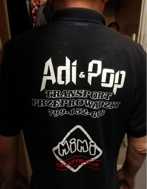 Tył czarnej koszulki polo z białym nadrukiem 'Adi & Pop Transport Przeprowadzki 799-152-616' oraz logo 'Mimi Reaktywacja' w dolnej części.