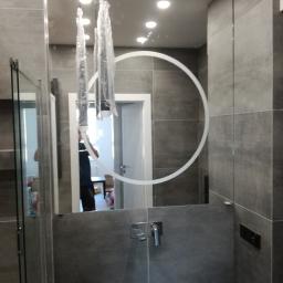 KWARC PRACOWNIA SZKLARSKA SPÓŁKA Z OGRANICZONĄ ODPOWIEDZIALNOŚCIĄ - Duże lustro łazienkowe z podświetlanym okręgiem, zamontowane na szarych płytkach, z widoczną armaturą i lampami w folii ochronnej.