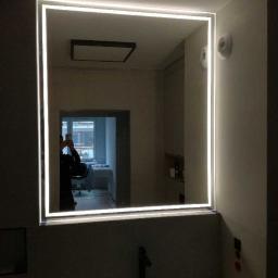 KWARC PRACOWNIA SZKLARSKA SPÓŁKA Z OGRANICZONĄ ODPOWIEDZIALNOŚCIĄ - Nowoczesne lustro łazienkowe z podświetleniem LED w kształcie kwadratu, odbijające fragment pomieszczenia z oknem i osobą robiącą zdjęcie telefonem.