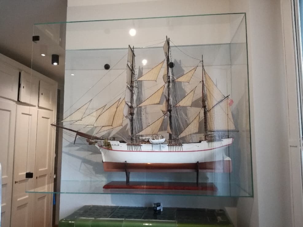 Model żaglowca 'Alicja' w szklanej gablocie, umieszczonej na szklanych półkach w pomieszczeniu z białą zabudową meblową.