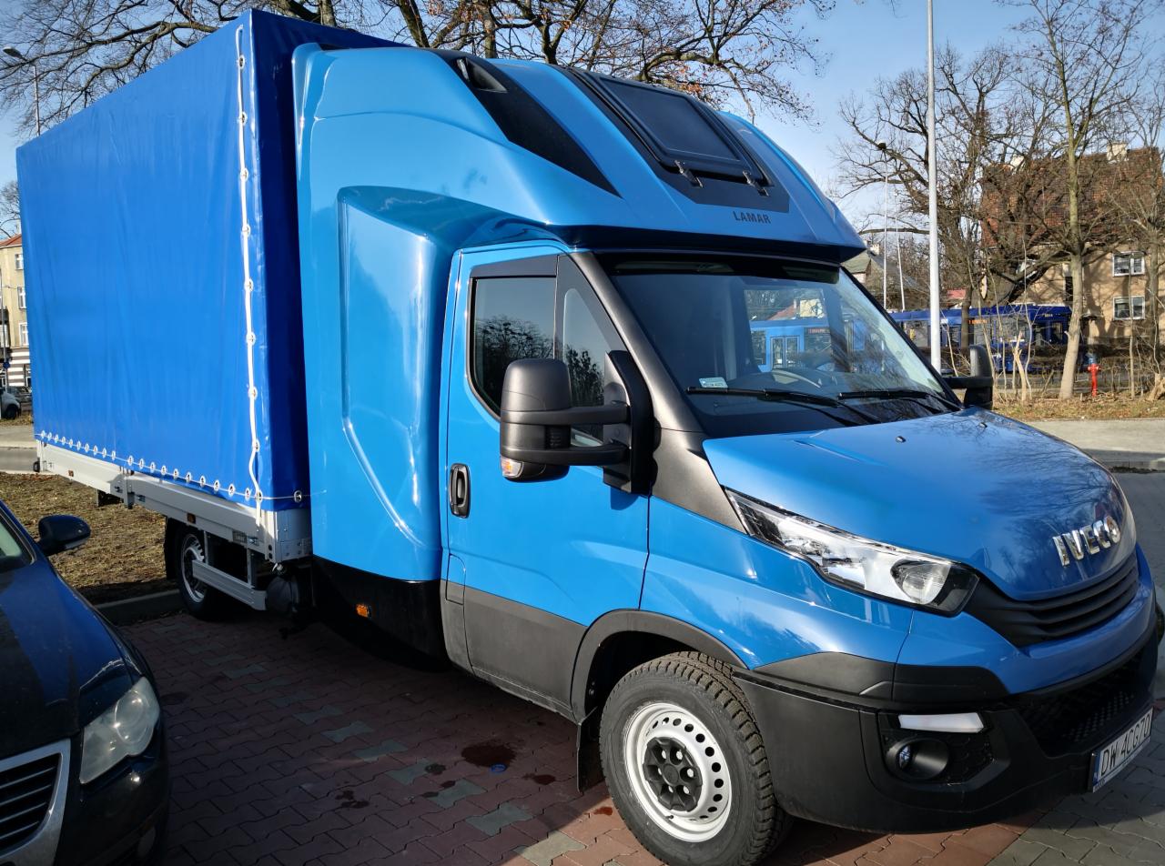 Niebieski samochód dostawczy z plandeką, marki Iveco, zaparkowany na brukowanej powierzchni, z widocznym napisem 'LAMAR' nad szybą przednią.