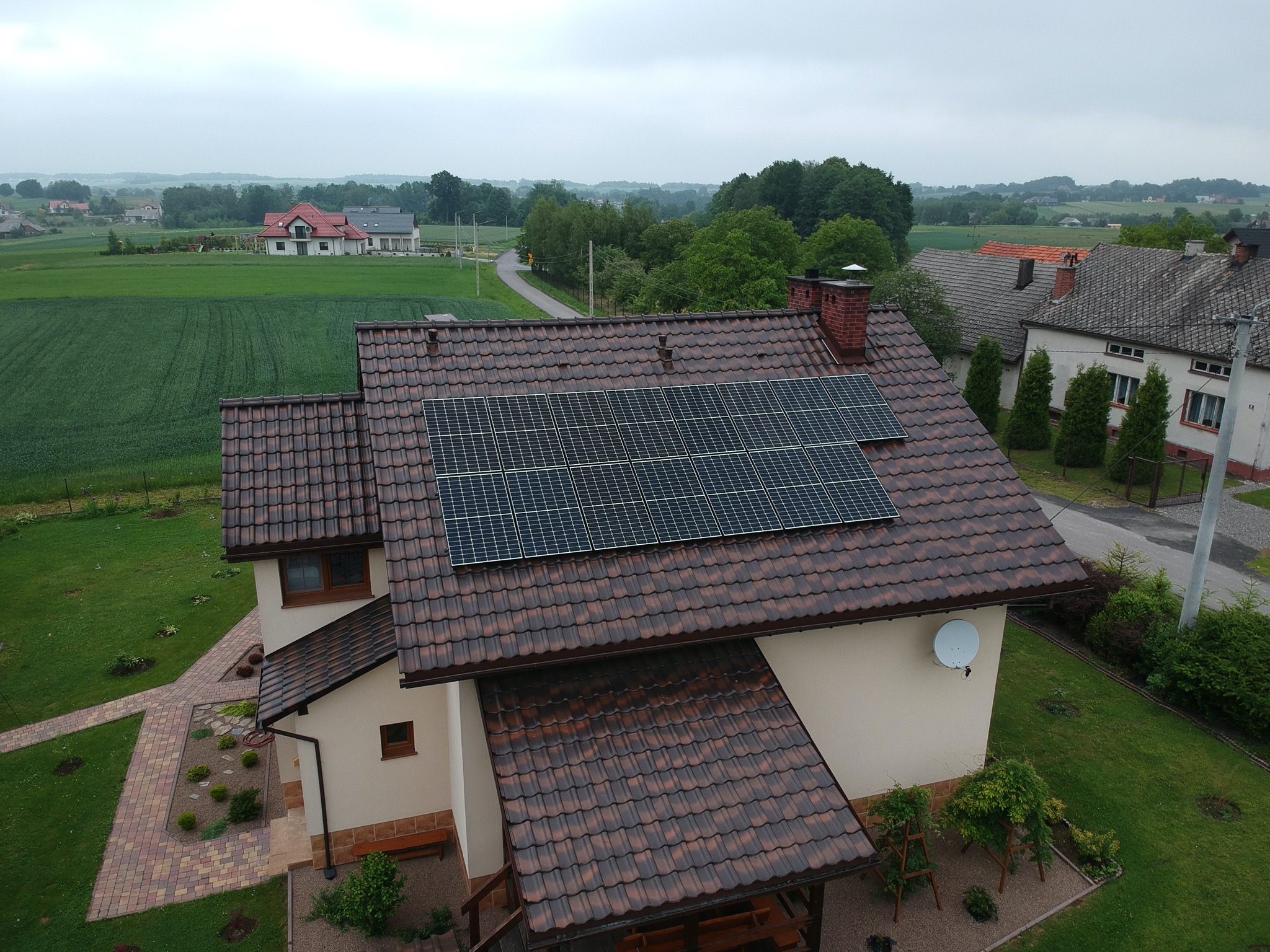 Widok z góry na dom z brązowym dachem pokrytym panelami słonecznymi, w tle wiejski krajobraz z polami i zabudowaniami.