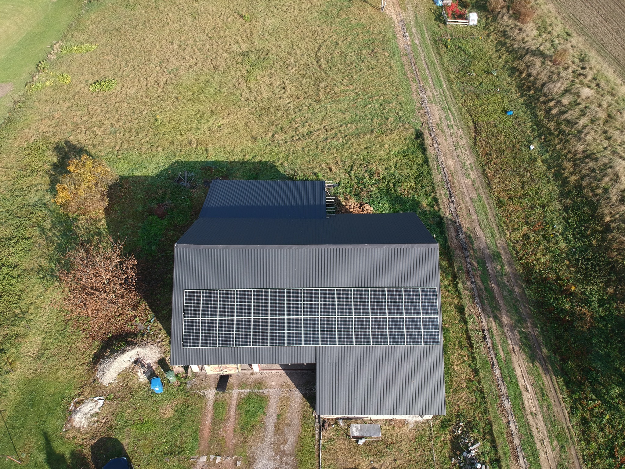 Widok z góry na budynek gospodarczy z czarnym dachem z blachy i instalacją paneli słonecznych, obok polna droga i drzewo z żółtymi liśćmi.