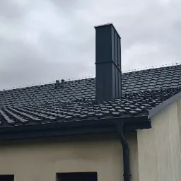 Szary dach z blachodachówki trapezowej i ciemny komin wentylacyjny na tle pochmurnego nieba. Widoczne obróbki blacharskie i rynna.