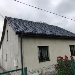Dom z nowym, ciemnym dachem z blachy trapezowej i czarnymi rynnami. Widoczne detale obróbek blacharskich. W tle pochmurne niebo i częściowo widoczni ludzie.