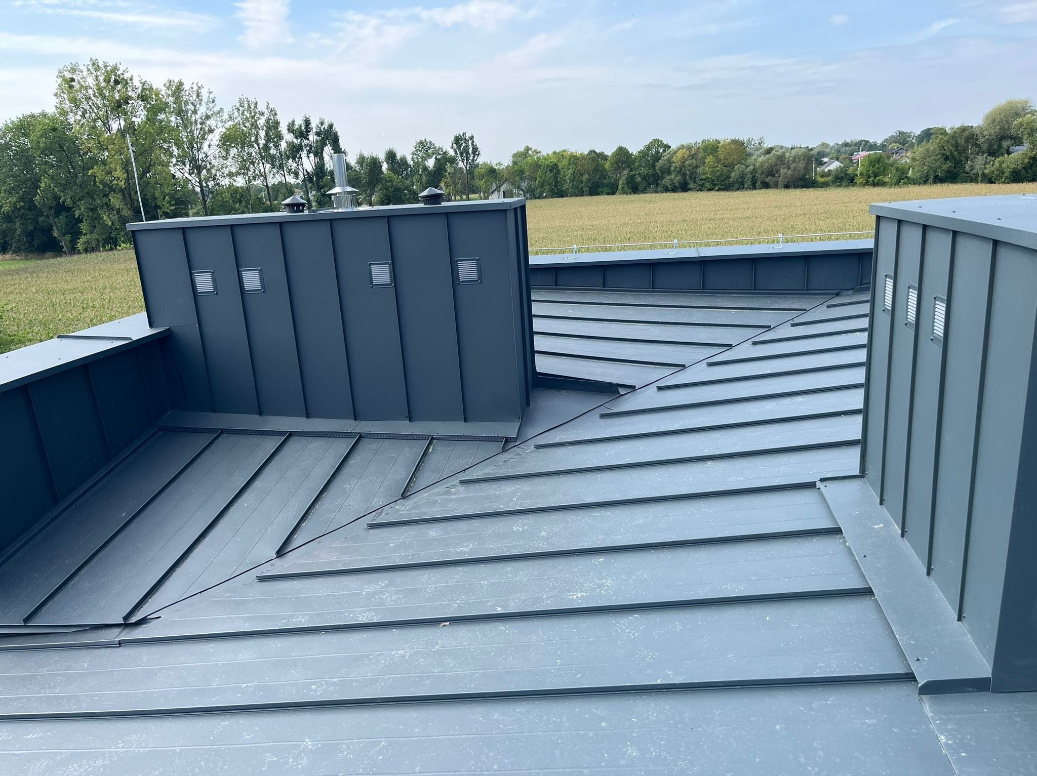 Szary dach z blachy na tle zieleni i błękitnego nieba. Widoczne kominy wentylacyjne i precyzyjne obróbki blacharskie. Nowoczesne krycie dachu.