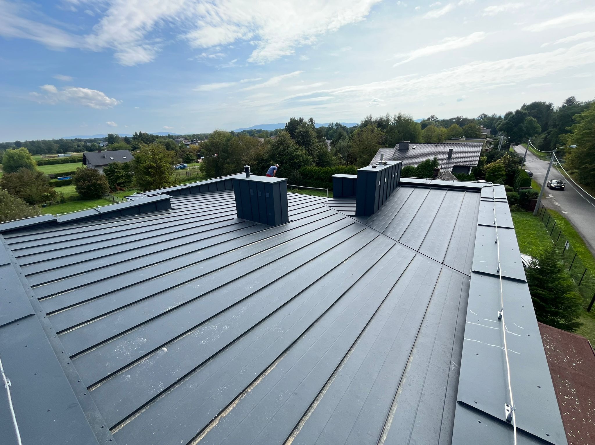 Szary, blaszany dach z kominami na tle zieleni i błękitnego nieba. Widoczny pracownik w oddali. Dach po kryciu, z wyraźnymi łączeniami blachy.