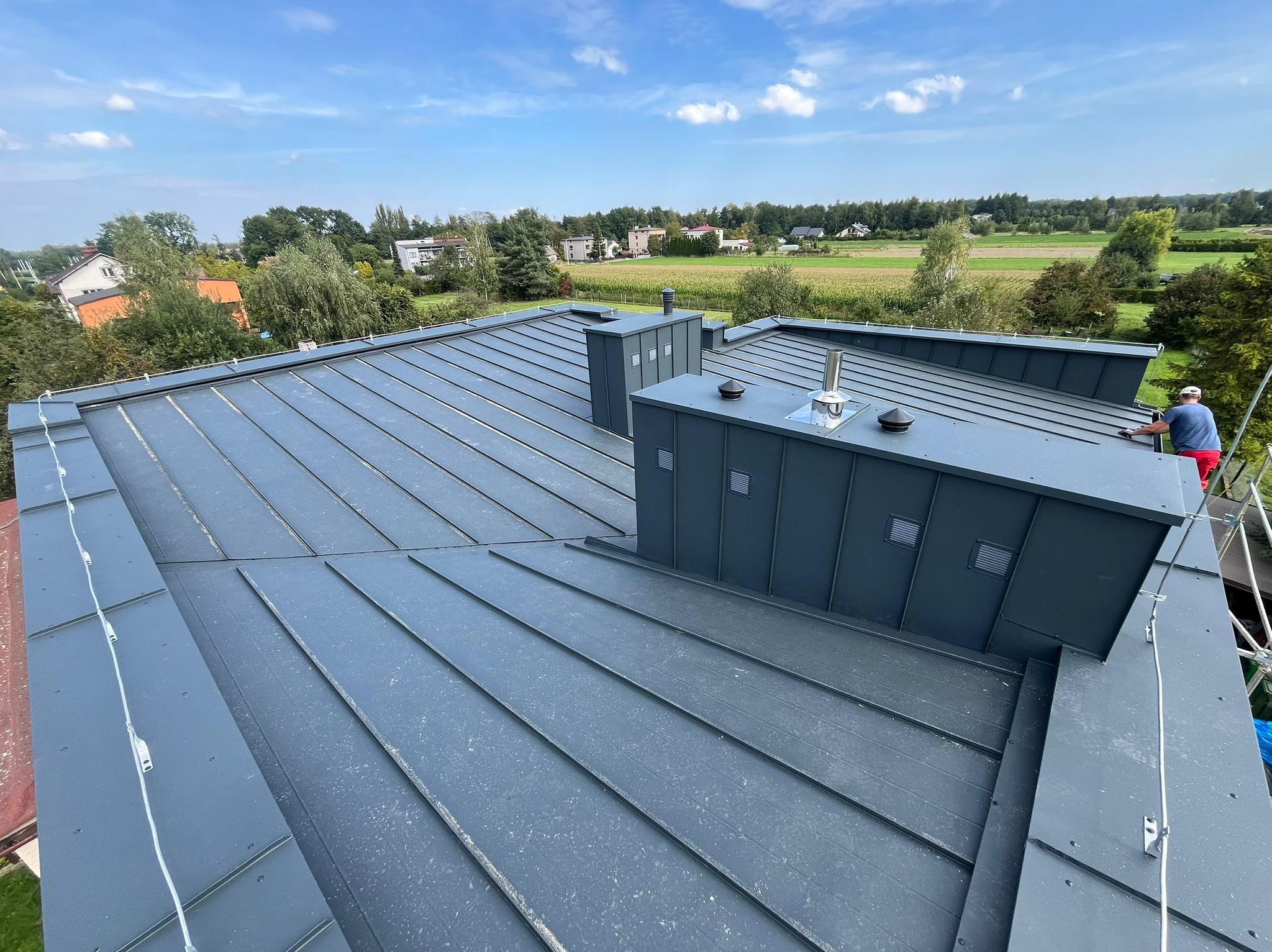 Szary, blaszany dach z kominami wentylacyjnymi na tle zielonych drzew i błękitnego nieba. Dekarz w trakcie pracy na dachu. Widok z góry.