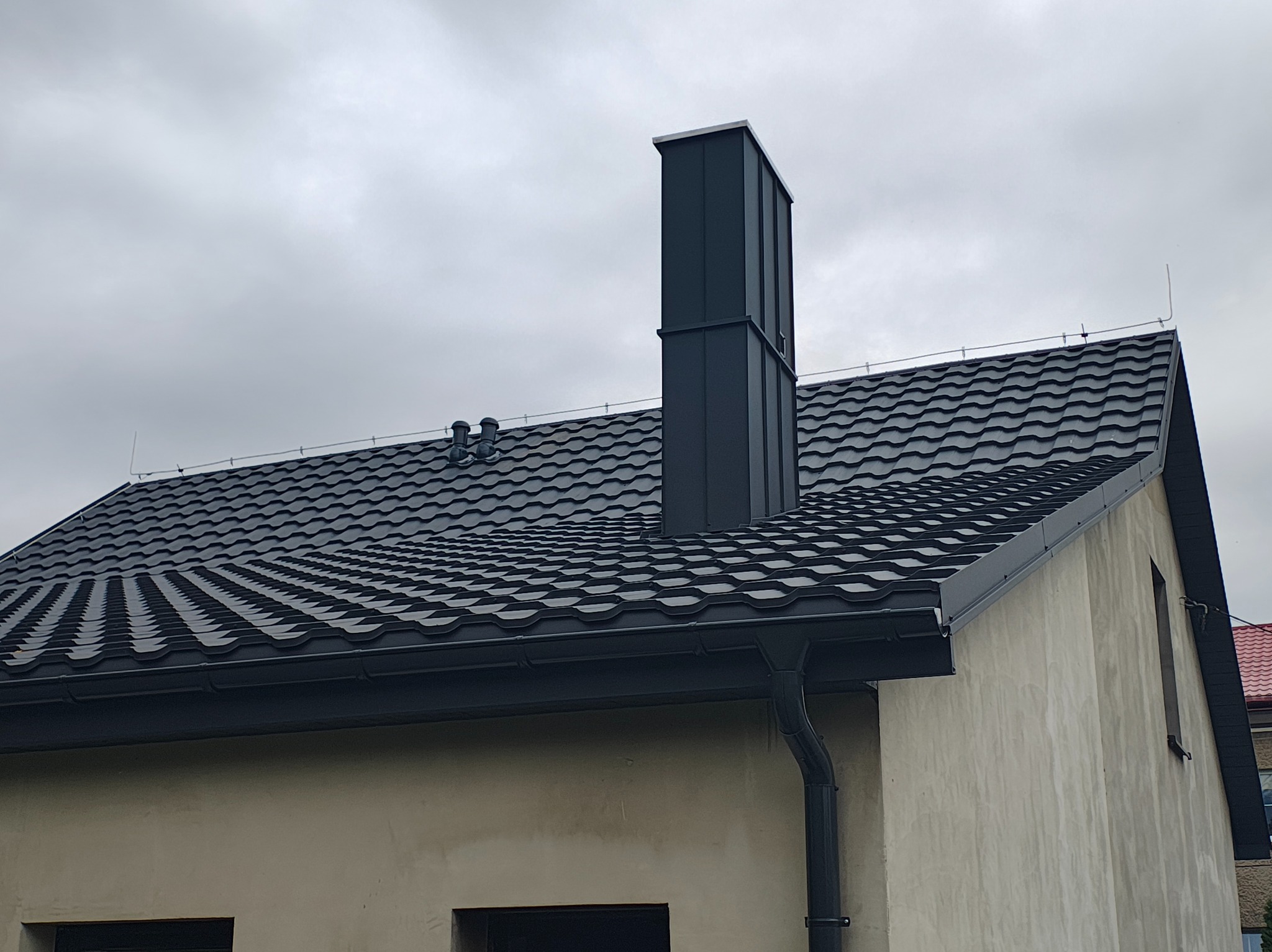 Szary dach z blachodachówki trapezowej i ciemny komin wentylacyjny na tle pochmurnego nieba. Widoczne obróbki blacharskie i rynna.