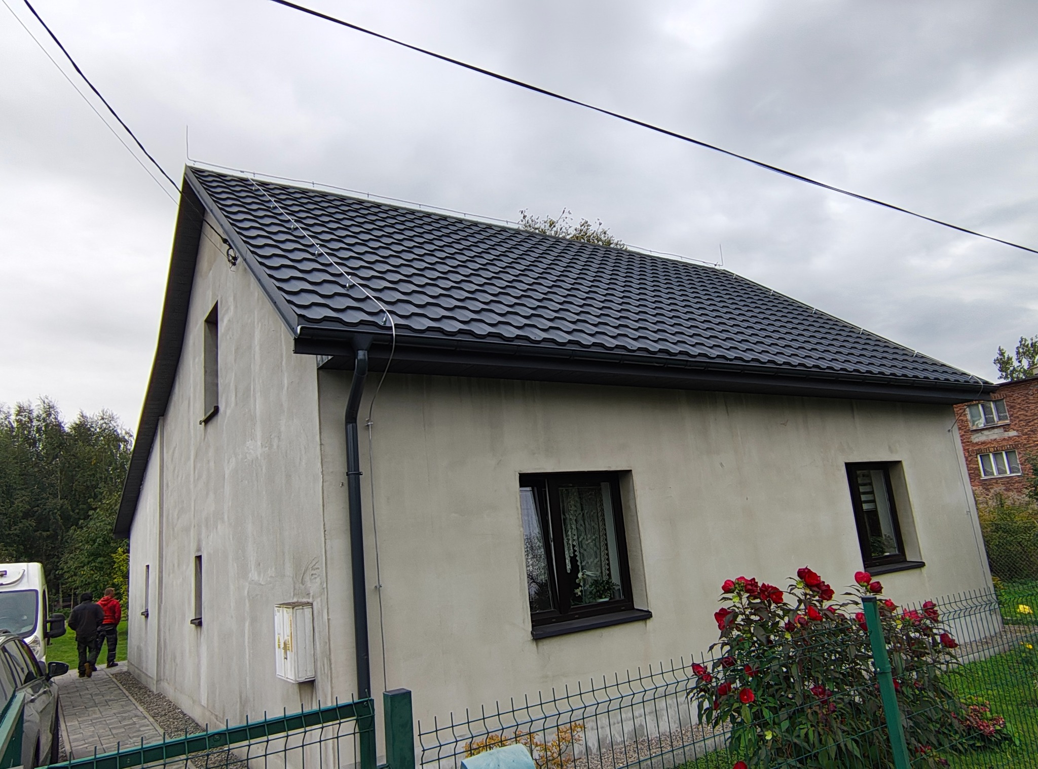 Dom z nowym, ciemnym dachem z blachy trapezowej i czarnymi rynnami. Widoczne detale obróbek blacharskich. W tle pochmurne niebo i częściowo widoczni ludzie.