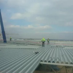 Montaż blachy trapezowej na dachu hali przemysłowej, widoczni pracownicy w kamizelkach odblaskowych i kaskach, w tle żuraw budowlany.