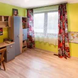 Pokój dziecięcy z zielonymi ścianami, drewnianą podłogą i meblami w odcieniach szarości i niebieskiego. Okno zasłonięte wzorzystymi zasłonami w czerwono-czarne szachownice. Na ścianie mapa świata...