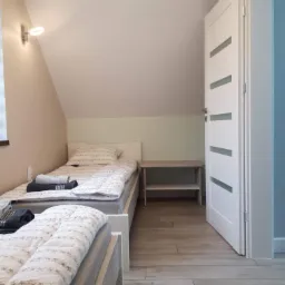 Pokój dwuosobowy na poddaszu z drewnopodobną podłogą, białymi łóżkami i drzwiami z przeszkleniami.