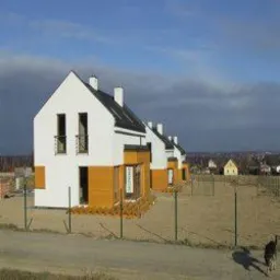 Rząd nowoczesnych domów murowanych z białymi ścianami i elementami z paneli w kolorze miodowym, widocznych z zewnątrz, ogrodzonych niskim płotem z siatki, na tle krajobrazu z zabudowaniami.