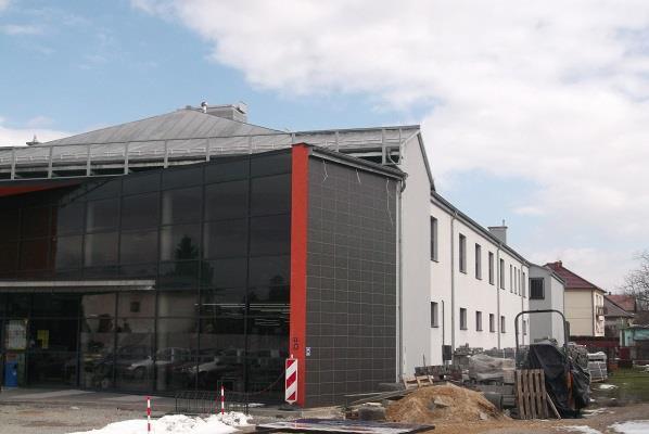 Nowoczesny budynek z dużą przeszkloną fasadą i elementami wykończenia z szarej płytki oraz czerwoną kolumną, widoczne materiały budowlane przed budynkiem.