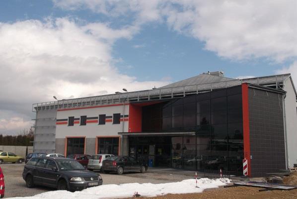 Nowoczesny budynek handlowo-usługowy z dużą przeszkloną fasadą, pomalowany w biało-szaro-pomarańczowe pasy, z parkingiem przed wejściem, częściowo pokrytym śniegiem.