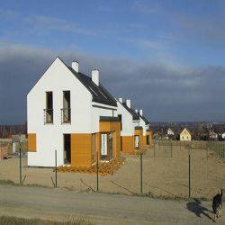 Rząd nowoczesnych domów murowanych z białymi ścianami i elementami z paneli w kolorze miodowym, widocznych z zewnątrz, ogrodzonych niskim płotem z siatki, na tle krajobrazu z zabudowaniami.